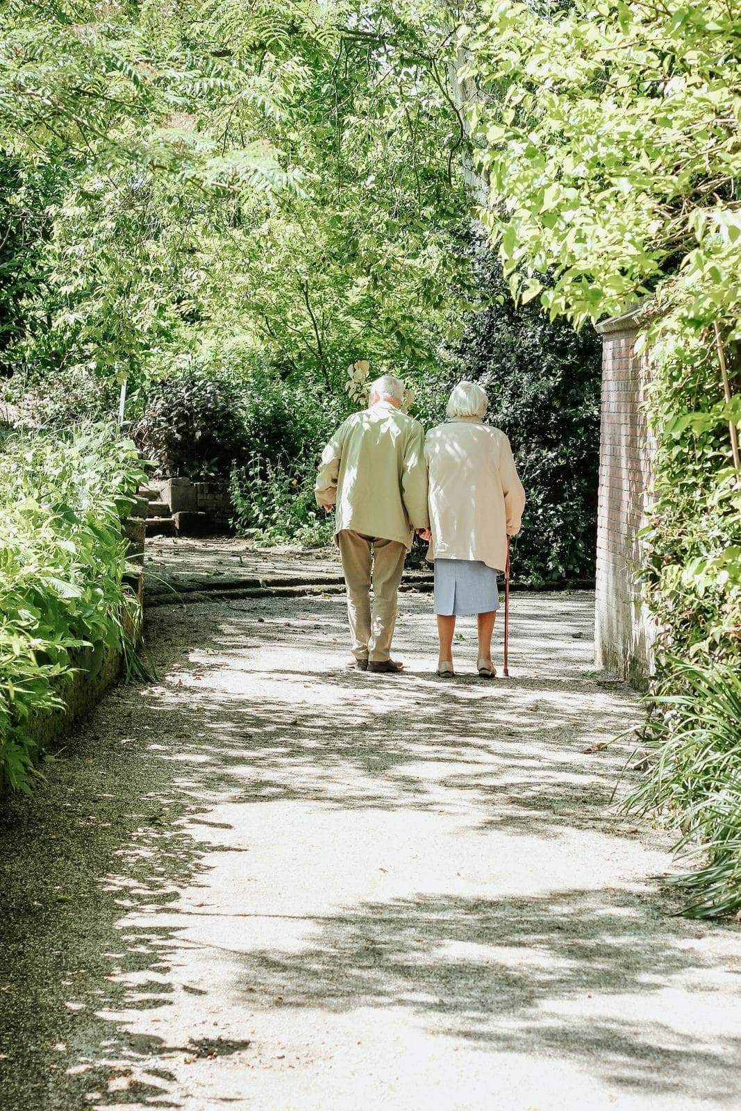 An older couple walking away down a shaded garden path, one of them using a cane.